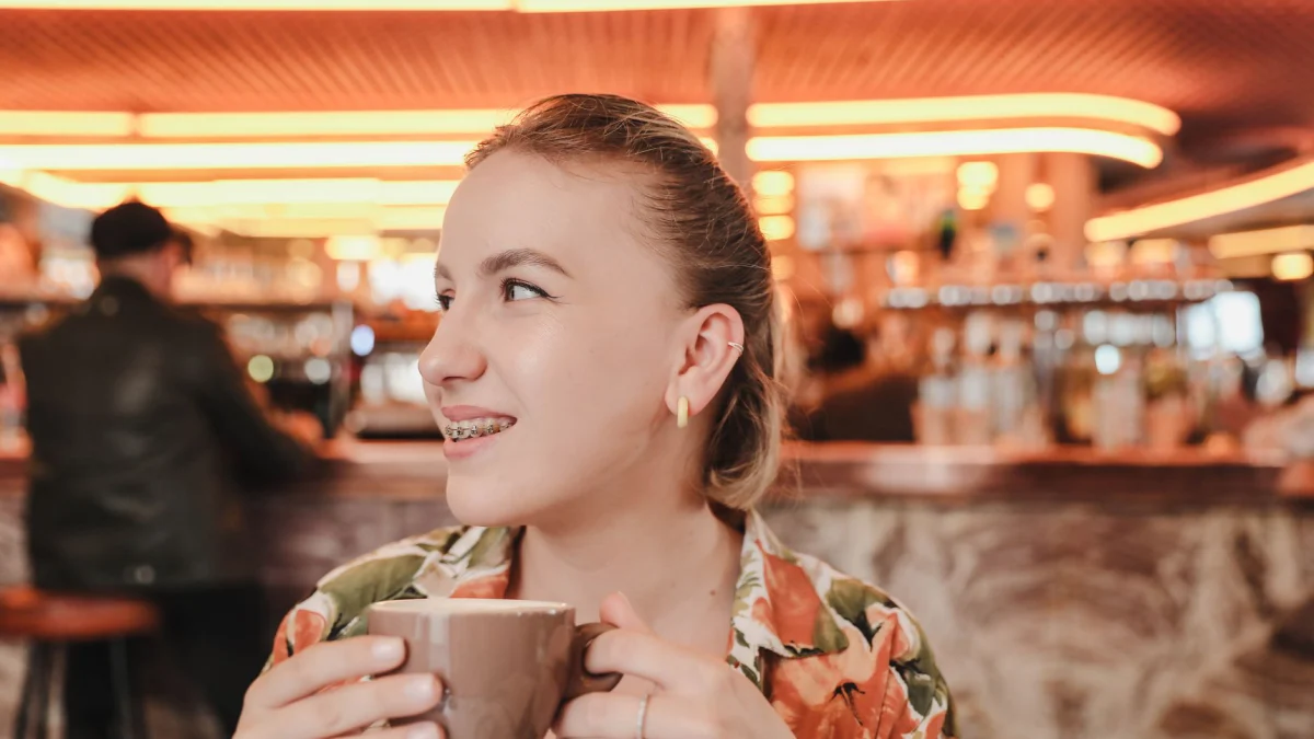 Portret van een vrolijke vrouw met een beugel die geniet van koffie in een stijlvolle cafésetting.