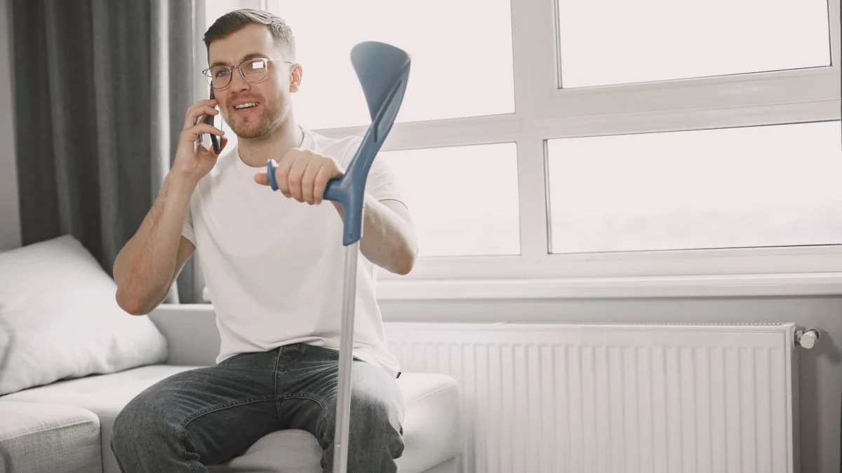 Volwassen man die op de bank zit, een kruk gebruikt en in een goed verlichte kamer aan de telefoon praat.