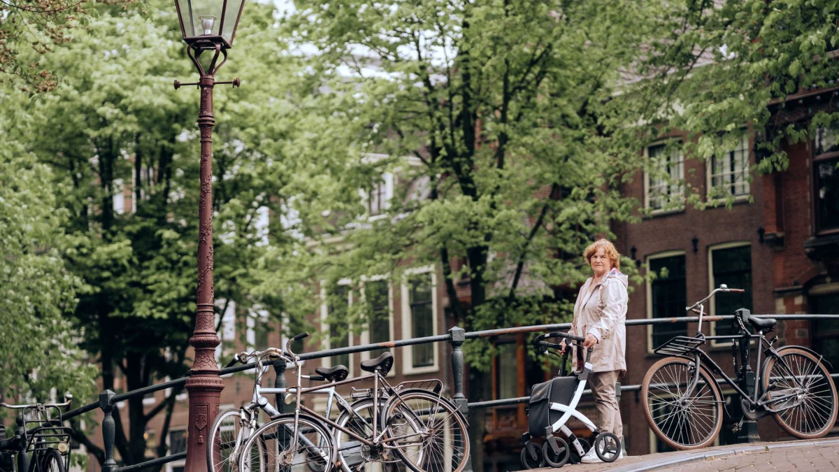 Oude vrouw die met een looprek op een schilderachtige brug in Amsterdam staat, omringd door fietsen en groen.