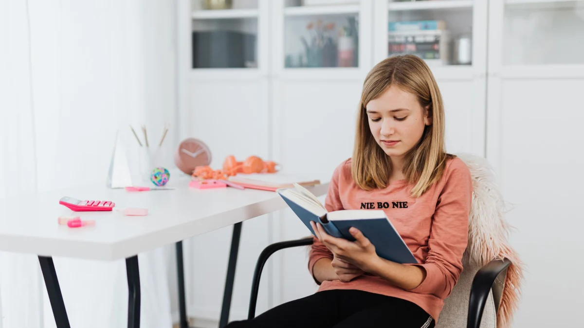 Tienermeisje in roze shirt leest boek in heldere, moderne ruimte voor studie of ontspanning.