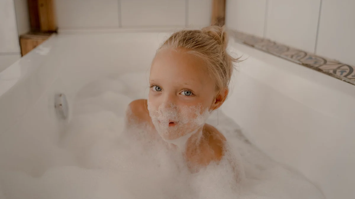 Een jong meisje met blond haar geniet van een speelse bubble bath in een witte badkuip binnenshuis.
