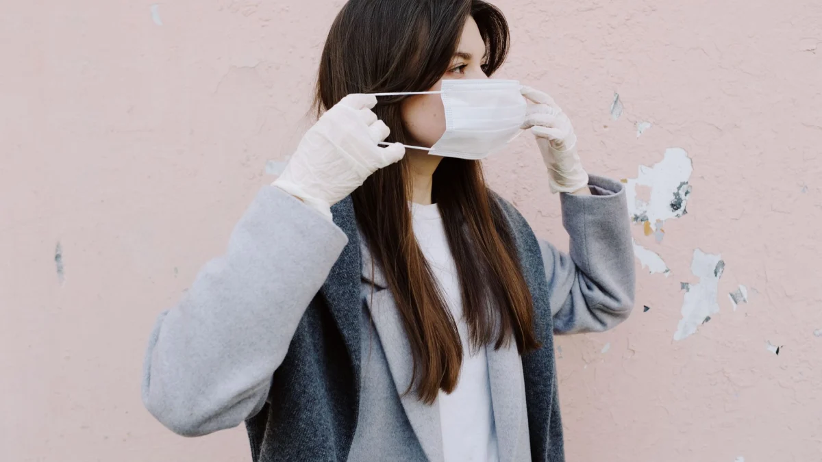 Jonge vrouw die een gezichtsmasker en handschoenen draagt voor veiligheid tijdens de pandemie. Zijaanzicht tegen een roze muur.