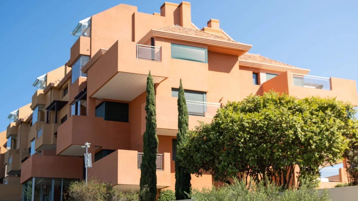 Een modern appartementengebouw in mediterrane stijl met terracottakleurige muren en weelderige vegetatie onder een heldere lucht.