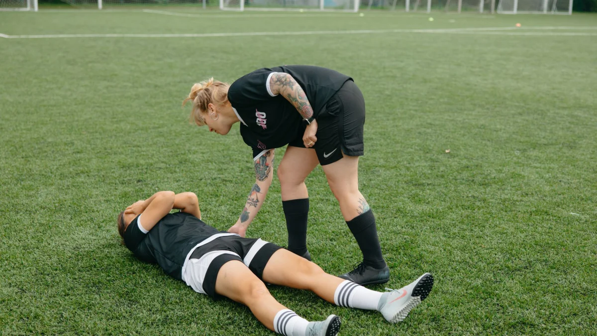 Een vrouwelijke voetballer helpt een geblesseerde teamgenoot op een groen voetbalveld tijdens een wedstrijd.
