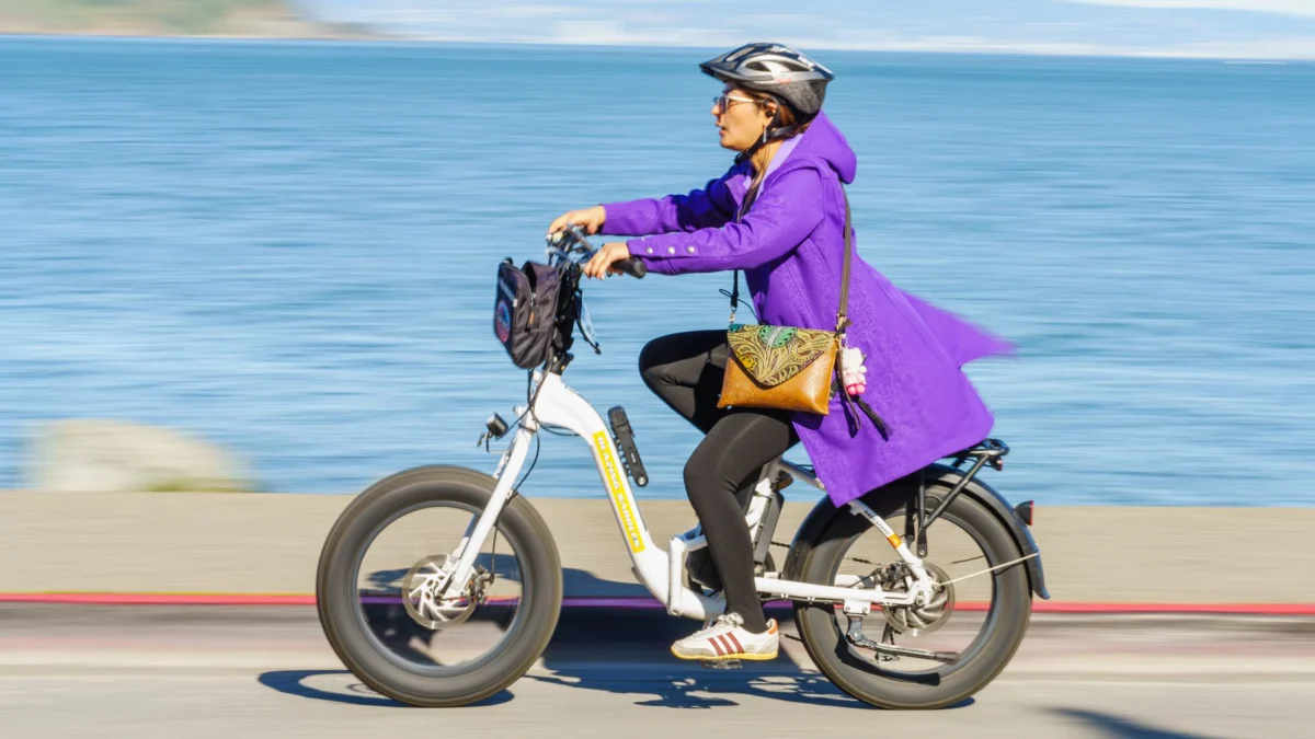 Op een zonnige dag fietst een vrouw op een elektrische fiets langs de waterkant, gekleed in een paarse jas en helm.