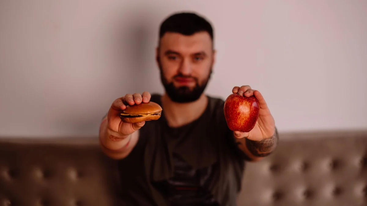 Een man houdt een appel en een cheeseburger vast, en toont de keuze tussen gezond en ongezond voedsel.
