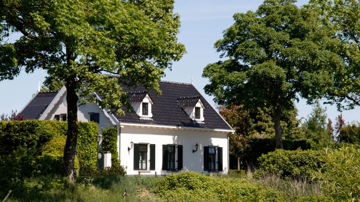 Idyllisch huisje op het platteland, omringd door groen en bomen onder een helderblauwe hemel.