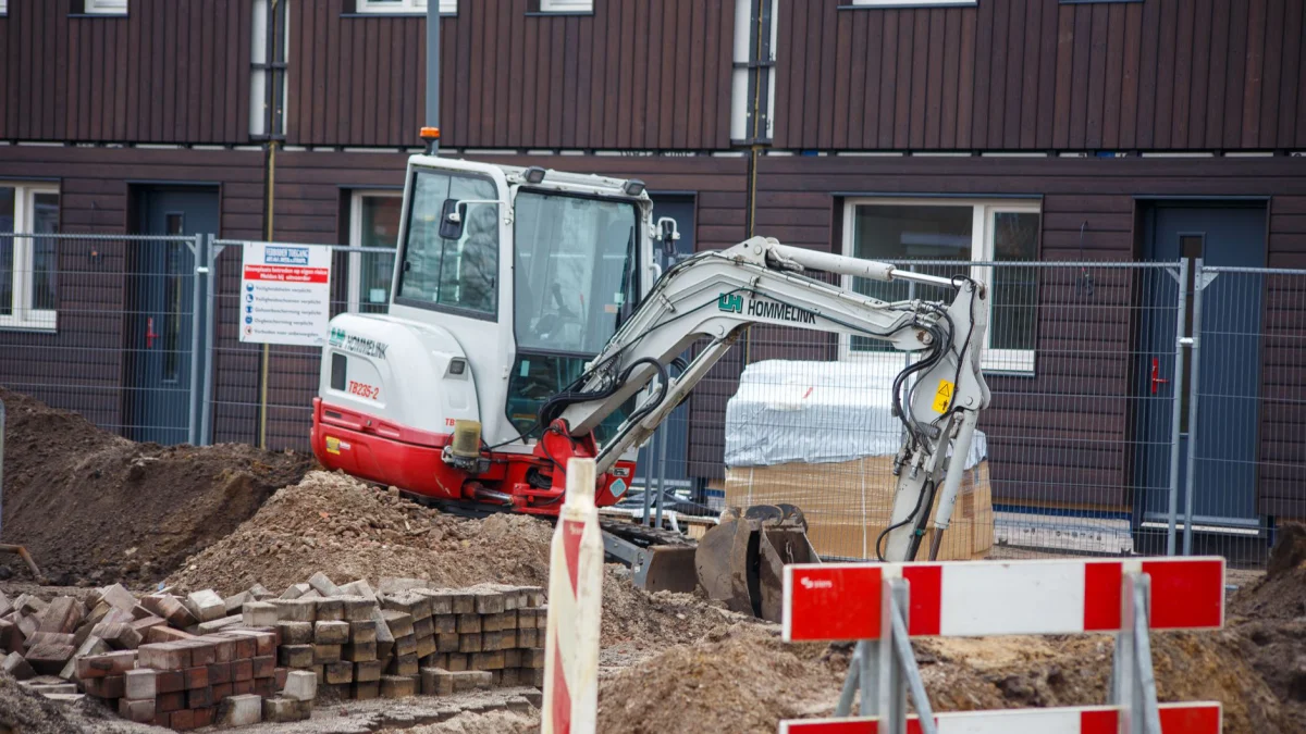 Een graafmachine die werkt op een bouwplaats in een woonwijk, omgeven door veiligheidshekken.
