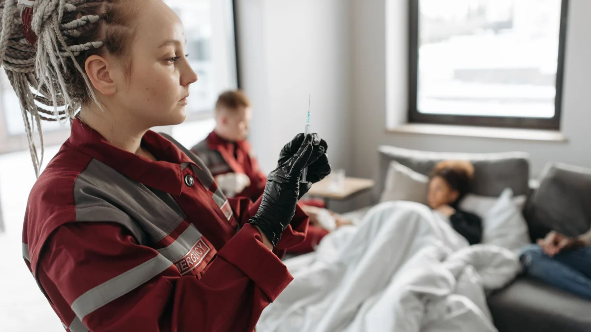 EHBO'er in een rode uniform bereidt een spuit voor een patiënt in een lichte ruimte.