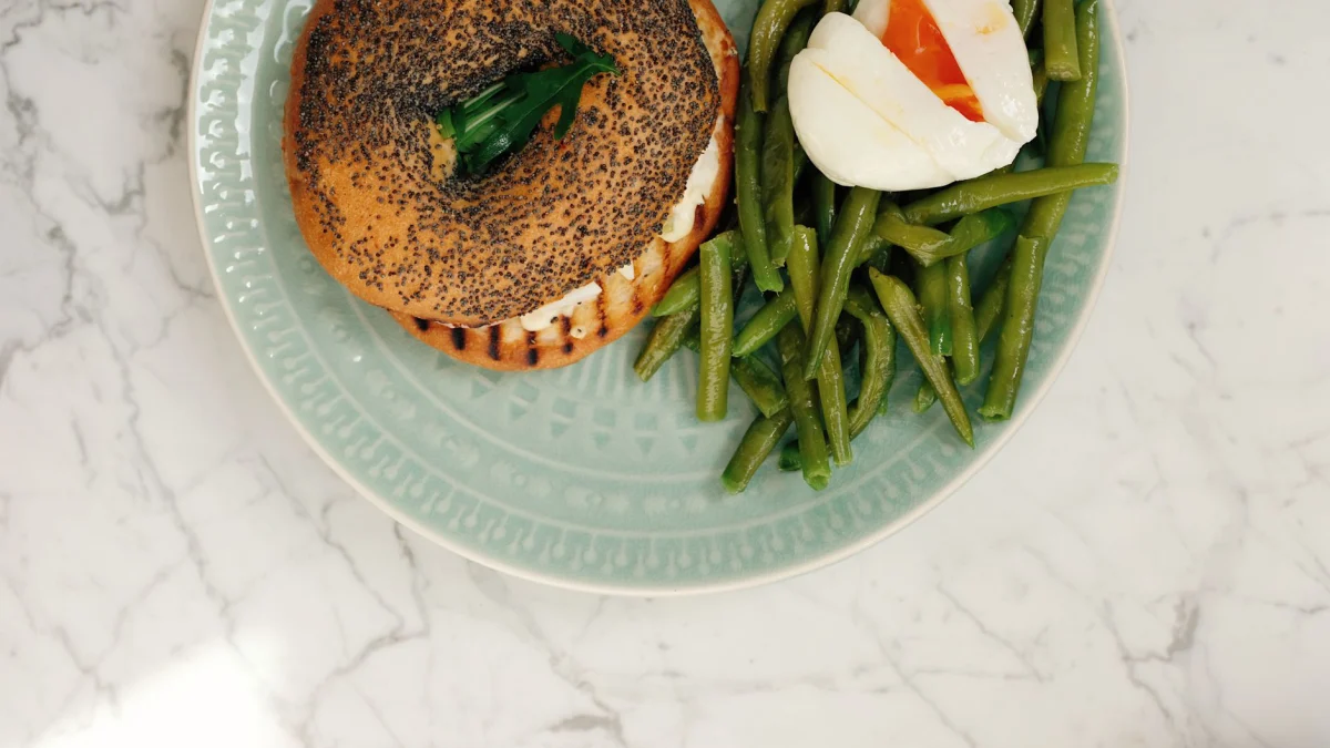 Een heerlijke en verse culinaire ontbijt met een poppy seed bagel met roomkaas, gepocheerd ei en groene bonen op een keramisch bord.