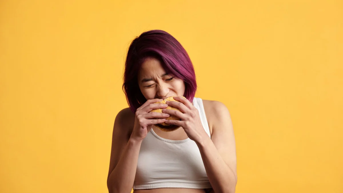 Een levendige studio-opname van een jonge vrouw met paarse haren die geniet van een hamburger tegen een gele achtergrond.