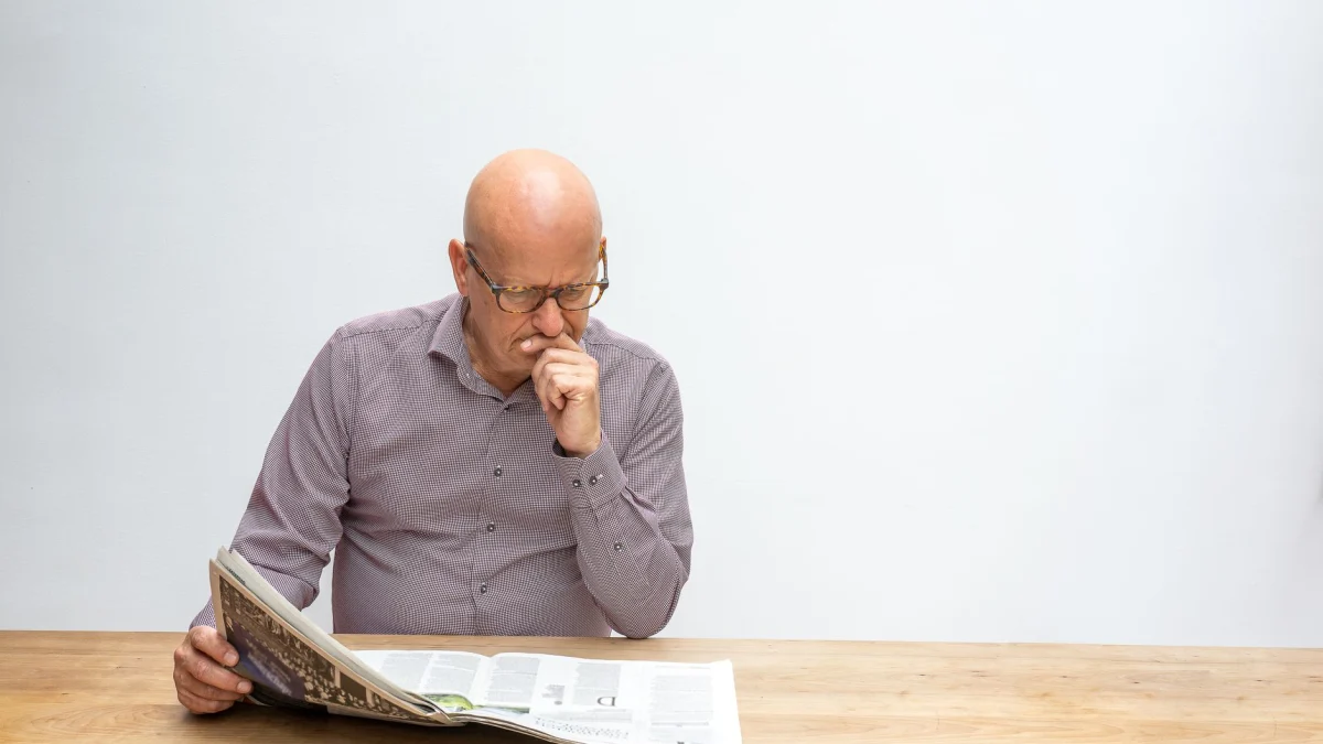 Oude man met een bril die een krant leest, zittend in gedachten aan een houten tafel op een witte achtergrond.