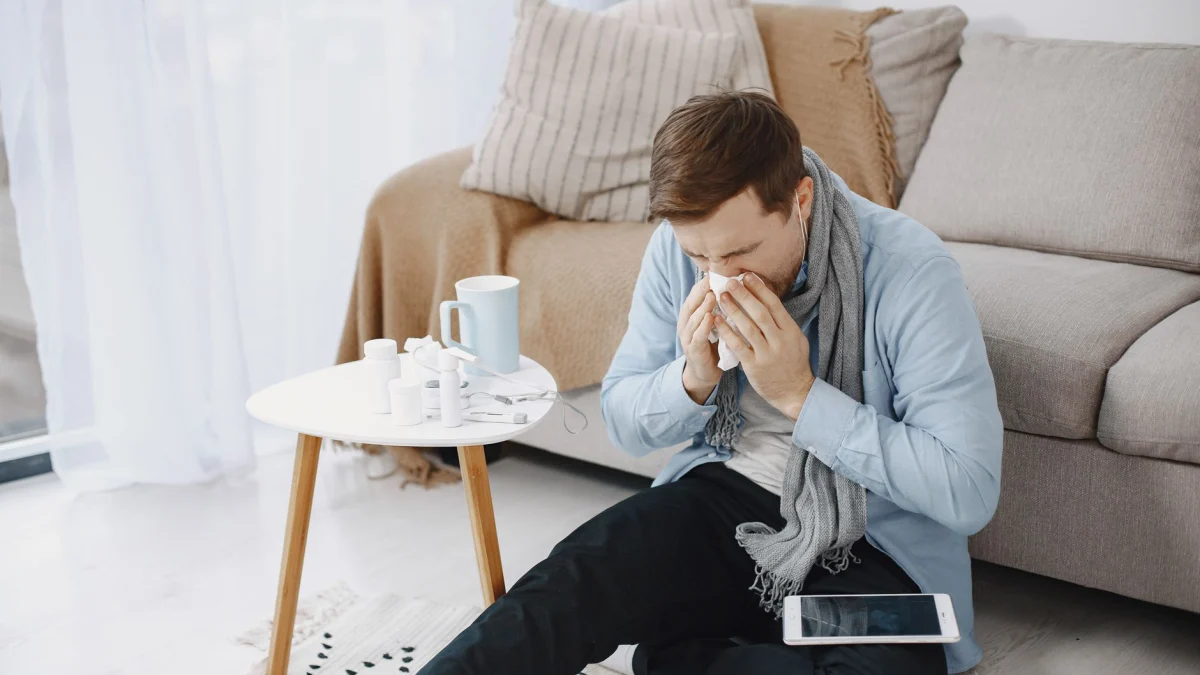 Een man die in een woonkamer in een tissue niest, omringd door een tafel, bank en digitaal tablet.