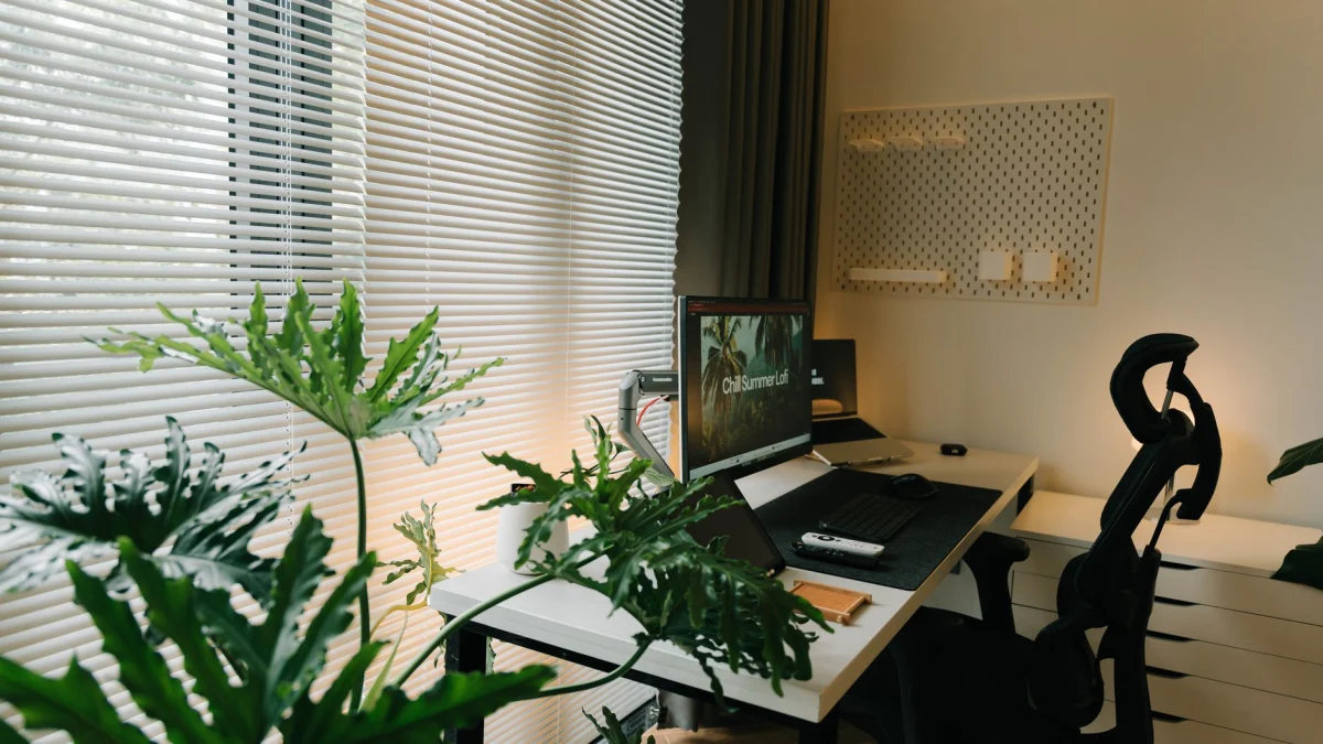 Een stijlvolle thuiswerkplek met een bureau, stoel en weelderige groene planten bij een raam met jaloezieën.