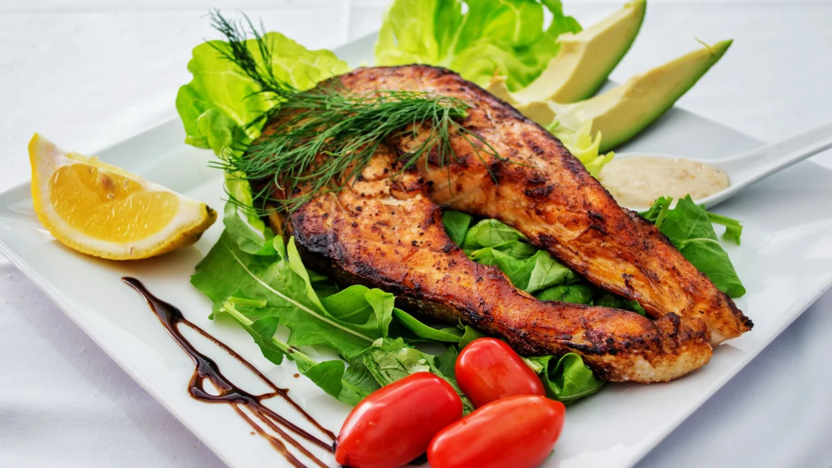 Heerlijke gegrilde zalmsteak geserveerd met avocado, sla, cherrytomaatjes en een schijfje citroen op een wit bord.