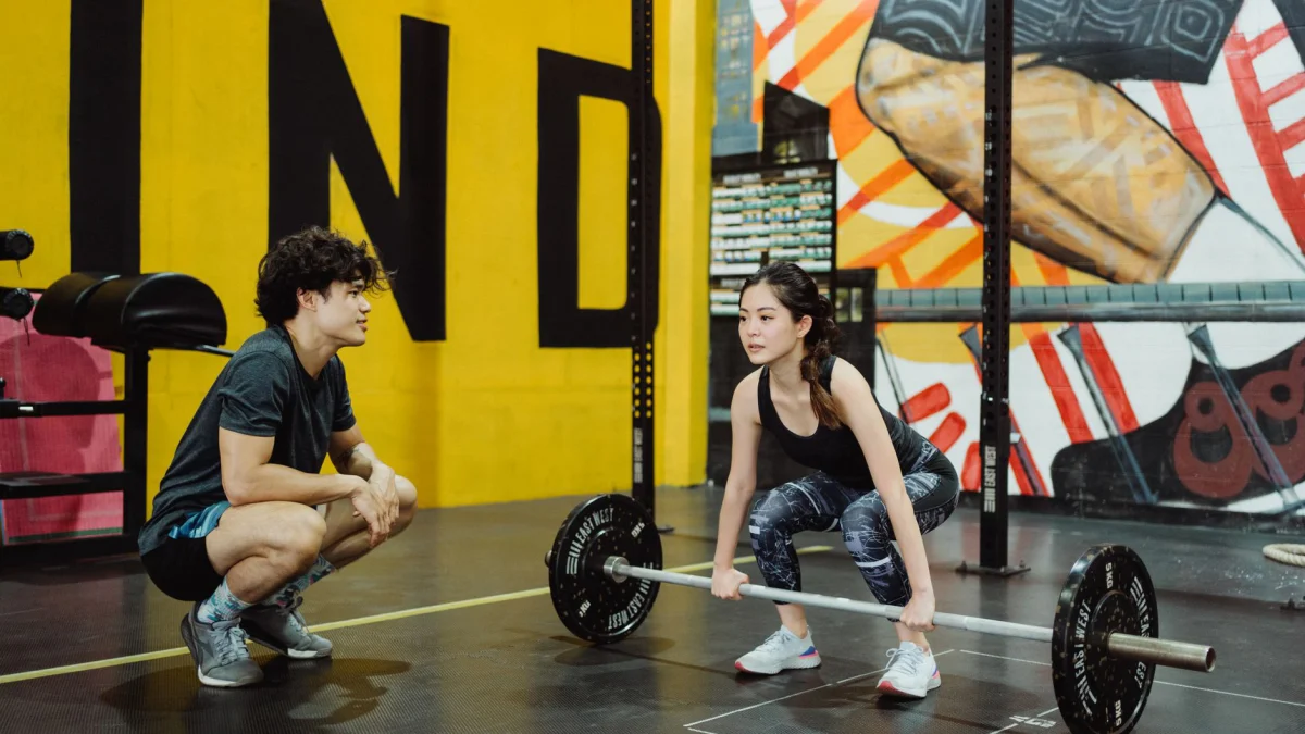 Een jonge vrouw tilt gewichten onder begeleiding van een trainer in een kleurrijke sportschool.