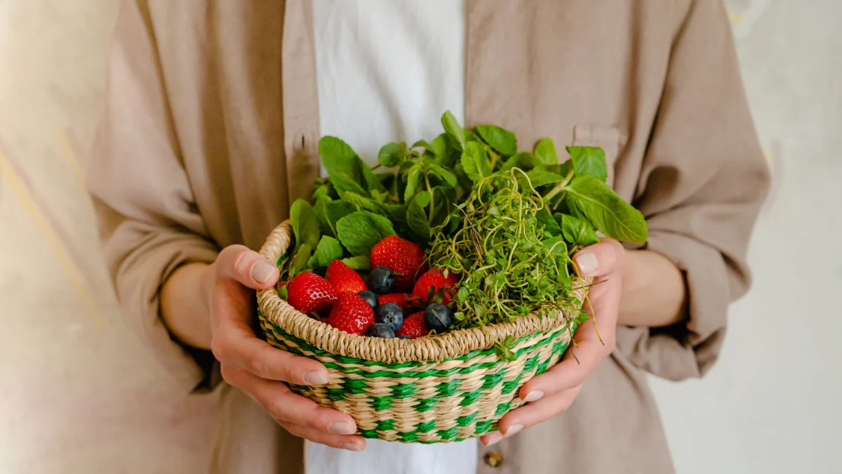 Close-up van een persoon die een mand vasthoudt met aardbeien, blauwe bessen en groenten.