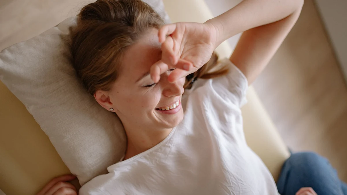 Glunderende vrouw in witte top ligt ontspannen op bed met hand voor haar gezicht en geniet van haar vrije tijd.