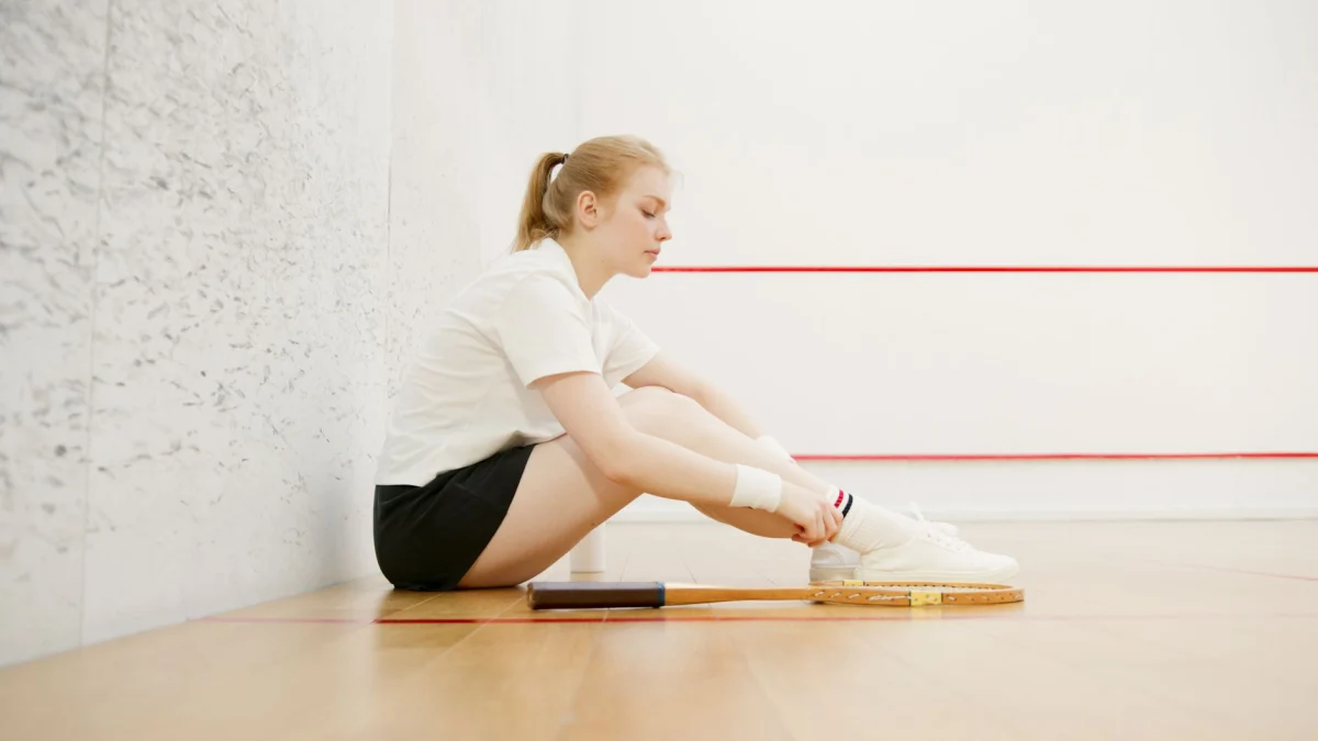 Zij-aanzicht van een jonge vrouw die op een squashbaan rust, gekleed in sportkleding en zittend met een racket.