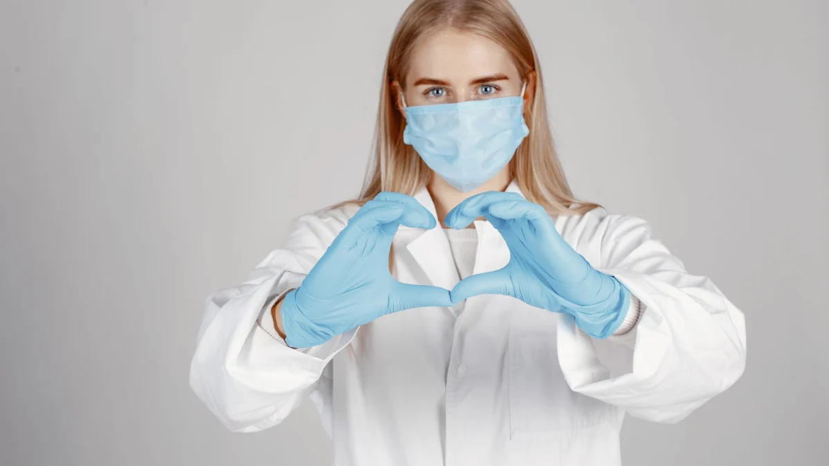 Zorgmedewerker met mondmasker en handschoenen maakt een hartvorm met haar handen, symbool voor zorg.