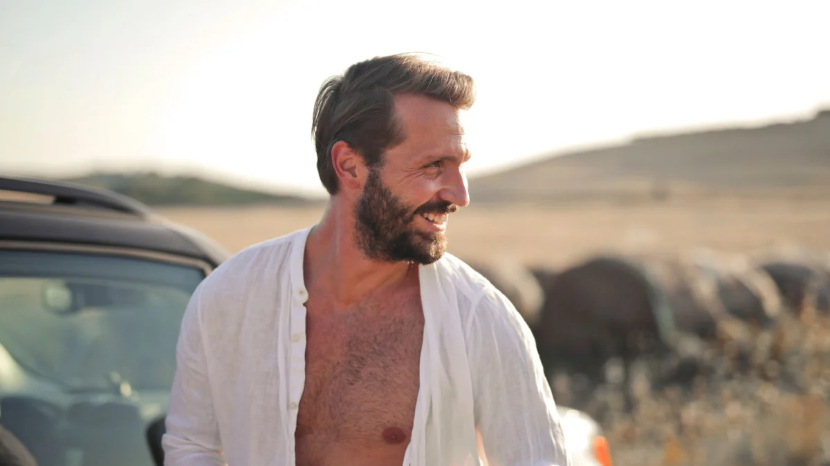 Een lachende man met een baard in een open shirt, genietend van een zonnige dag buiten naast een auto.