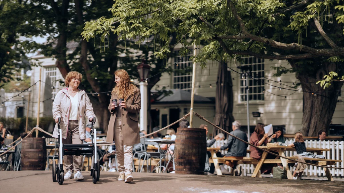 Oudere vrouw met een looprek en jonge metgezel die door een levendige buurt in Amsterdam wandelt.
