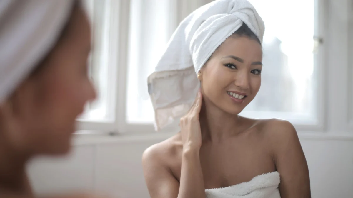 Een vrouw gewikkeld in een handdoek glimlacht naar haar reflectie in een heldere badkamer.